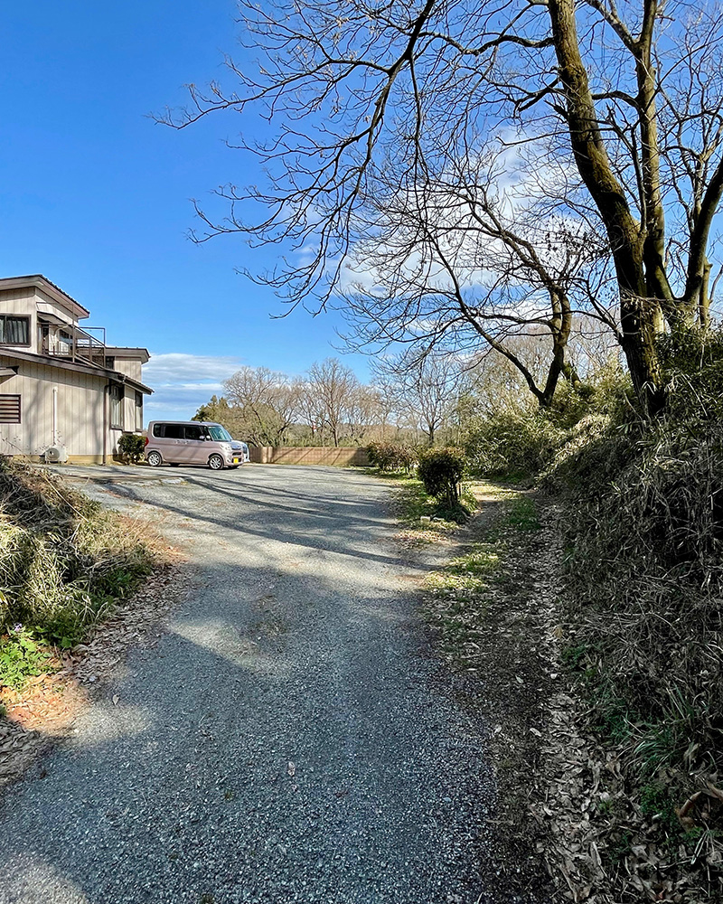 民家の敷地の右側から密かに街道が続く写真